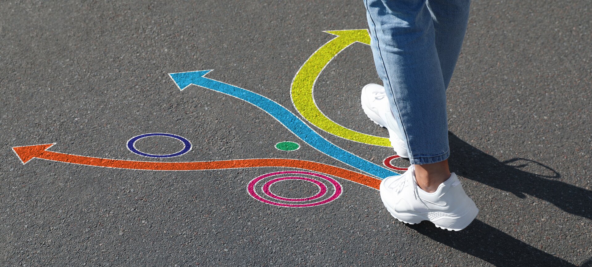Fotografia przedstawia nogi nieokreślonej postaci, które stoją na rozstaju dróg, w postaci kolorowych strzałek - pomarańczowej, niebieskiej, żółtej.