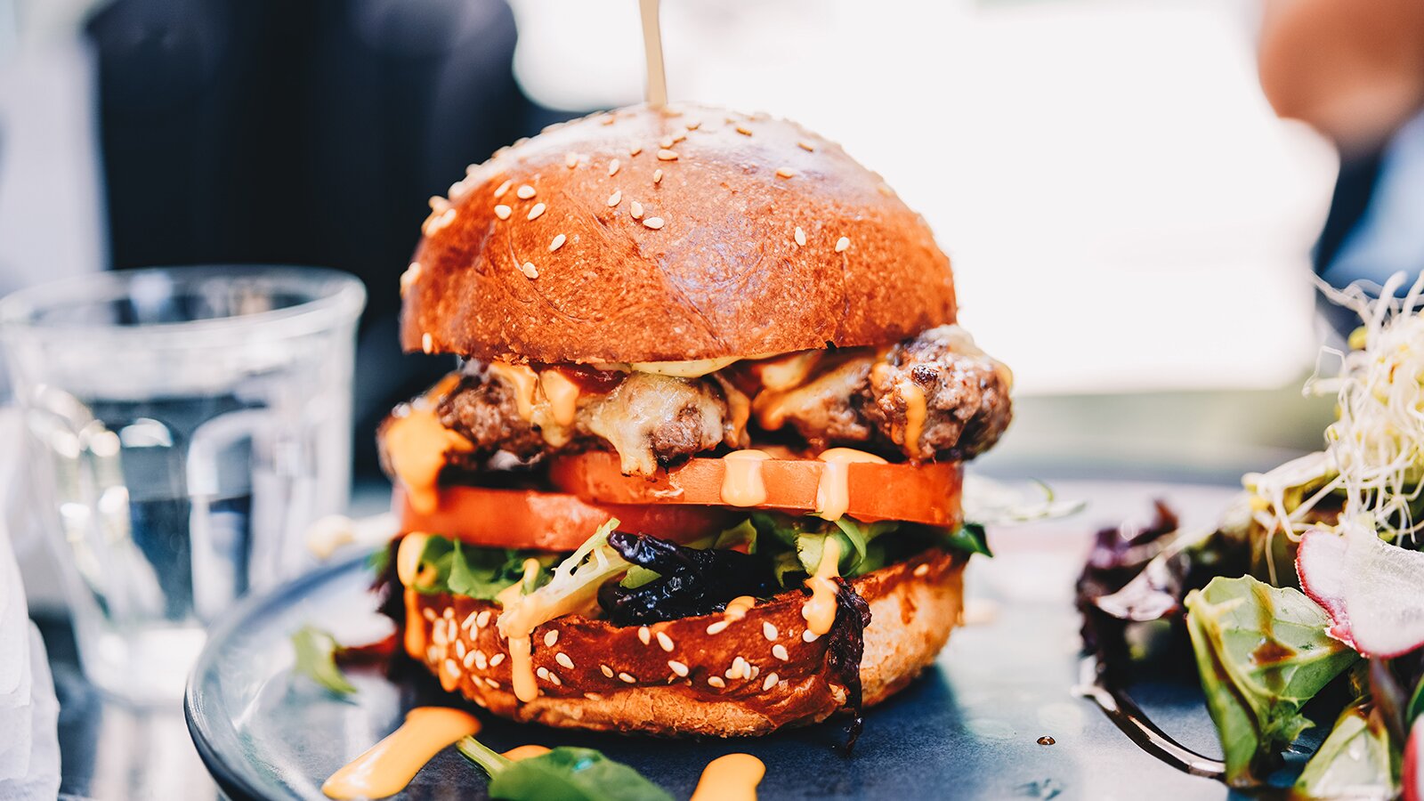 Zdjęcie przedstawia kanapkę typu cheeseburger z dużą ilością dodatków, bułkę posypano sezamem. Obok widoczna szklanka wody. 