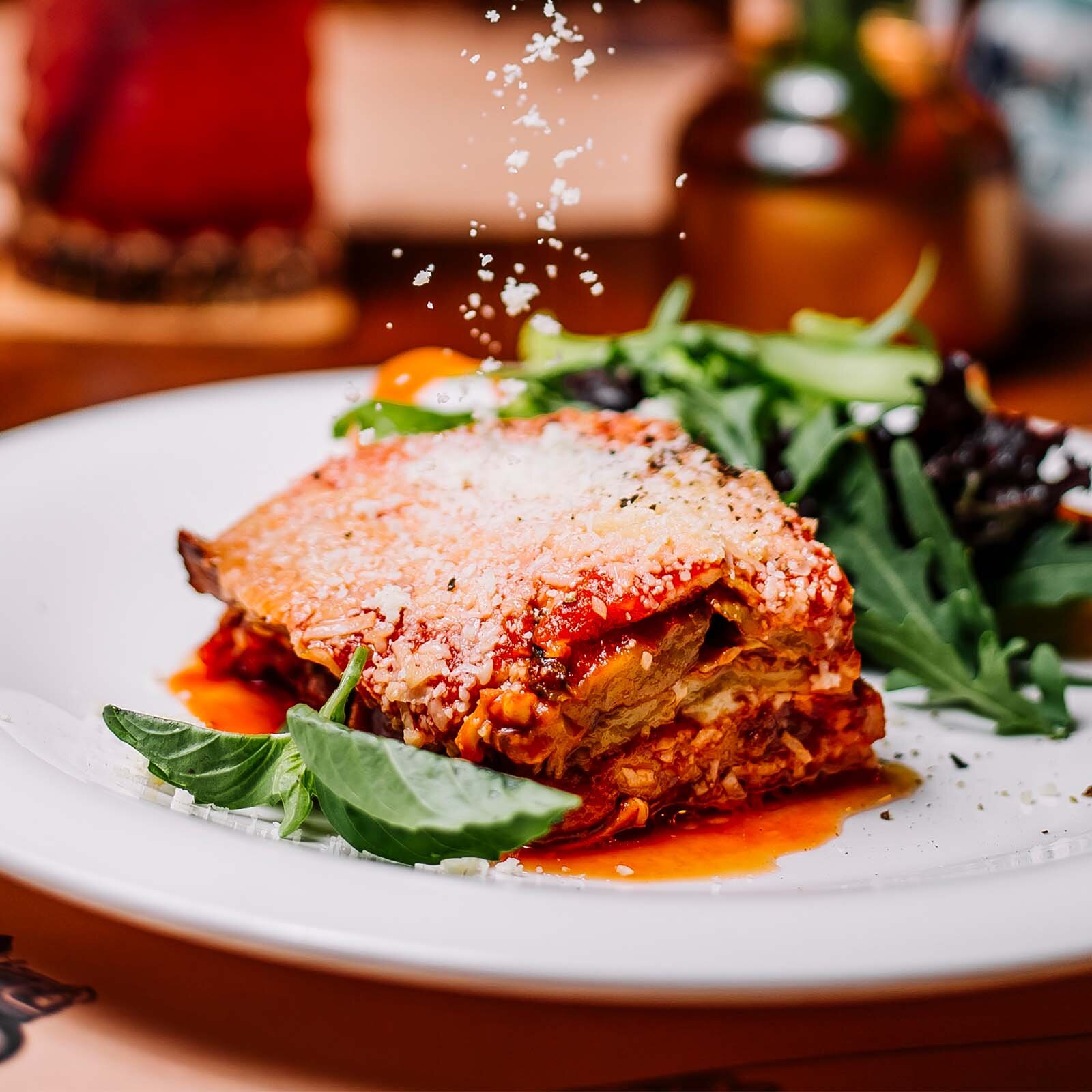 Zdjęcie przedstawia kawałek lasagne posypanej parmezanem i ułożonej na talerzu.