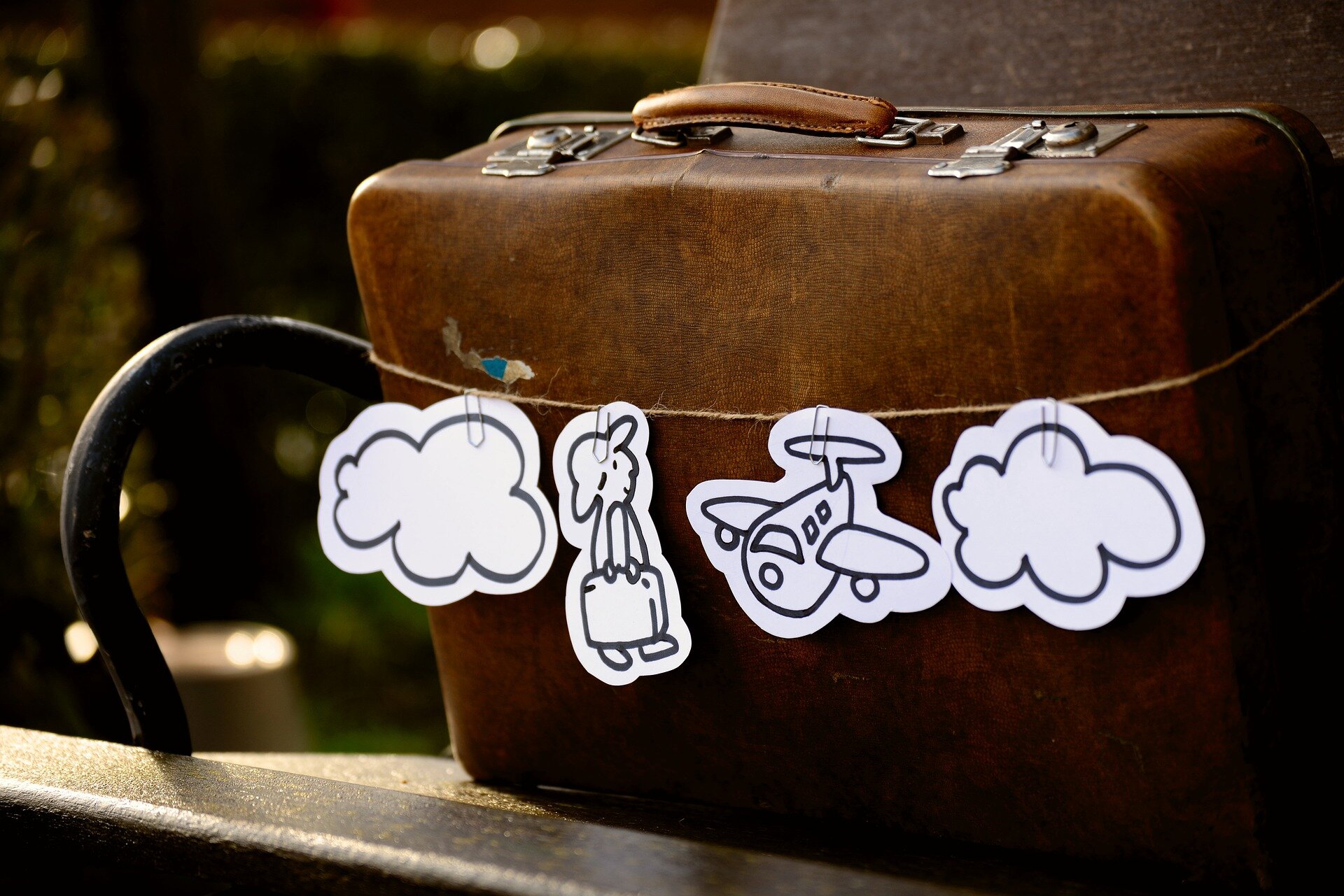 Zdjęcie przedstawia walizkę obwiązaną dookoła sznurkiem, na którym zawieszono 4 wycinanki. Dwie skrajne to chmury, a dwie środkowe to postać człowieka z walizką oraz samolot.