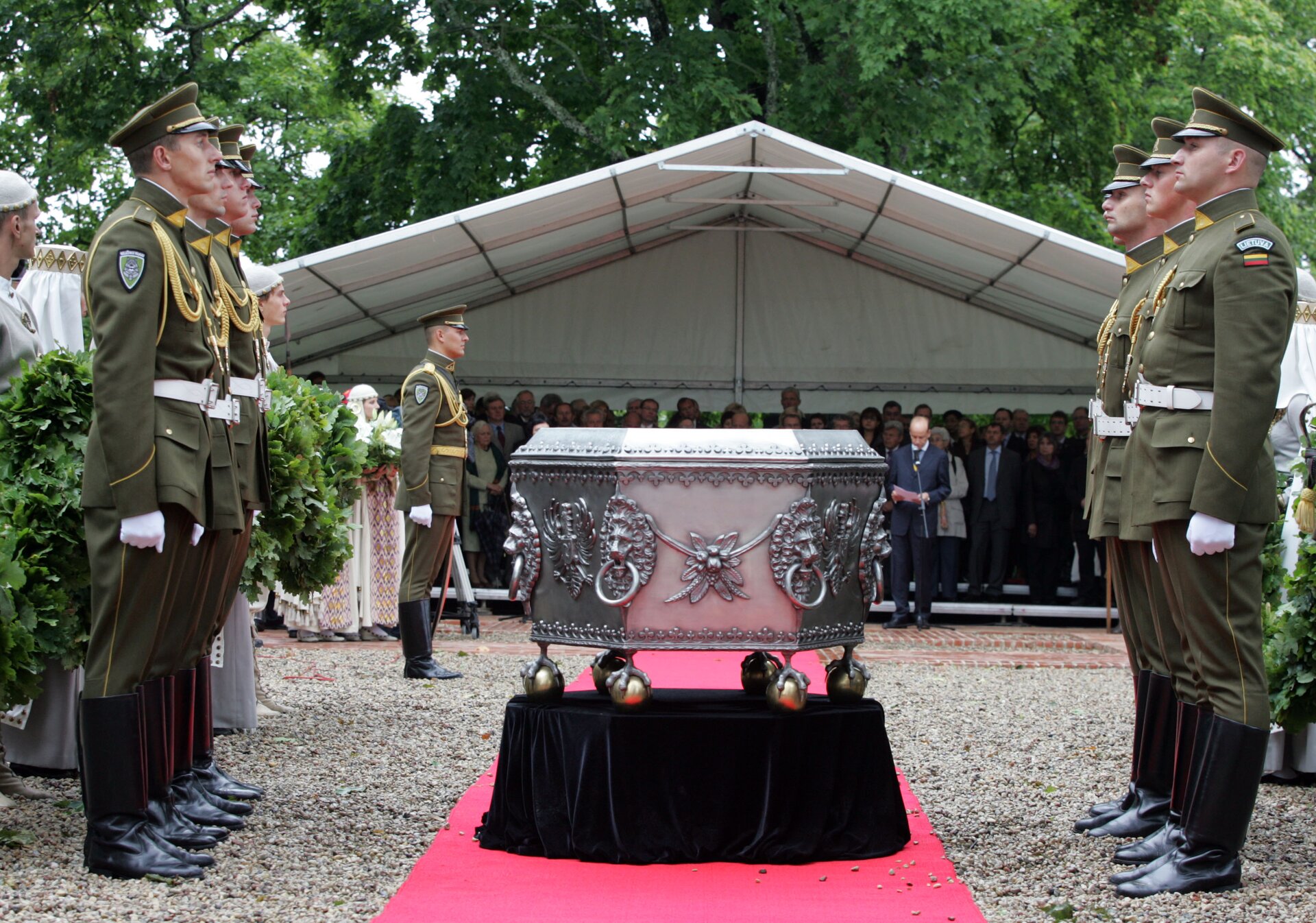 Zdjęcie przedstawia stojącą na środku urnę. Urna jest metalowa, ma kształt sześciokąta. Zdobią ją wizerunki lwów, którzy mają w okolicach ust i nosa powkładane koła. Na górze i na dole urnę oplata łańcuch z wizerunkiem małych krzyży. Urna jest ba sześciu okrągłych nóżkach, które mają na sobie wizerunek szponów potaków. Przy urnie stoją żołnierze (po trzech z każdej strony). W tle widać namiot i obecnych tam ludzi. 