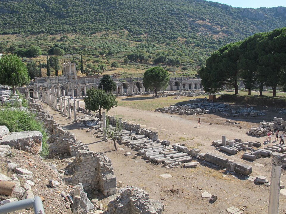 Zdjęcie przedstawia pozostałości zabudowań na planie kwadratu. Ruiny są na obrzeżach kwadratu, środek jest pusty. Widoczne są kolumny, które pierwotnie stały jedna przy drugiej w równych rzędach. Teraz większość leży na ziemi. W tle wzgórze porośnięte zielona roślinnością.