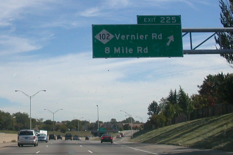 Na zdjęciu znajduje się trzypasmowa ulica, na której są samochody. W oddali widać zjazd w prawą stronę. Nad ulicą wisi od prawej strony tablica informacyjna z napisem Exit 225; 102 Vernier Road, 8 Mile Road. 