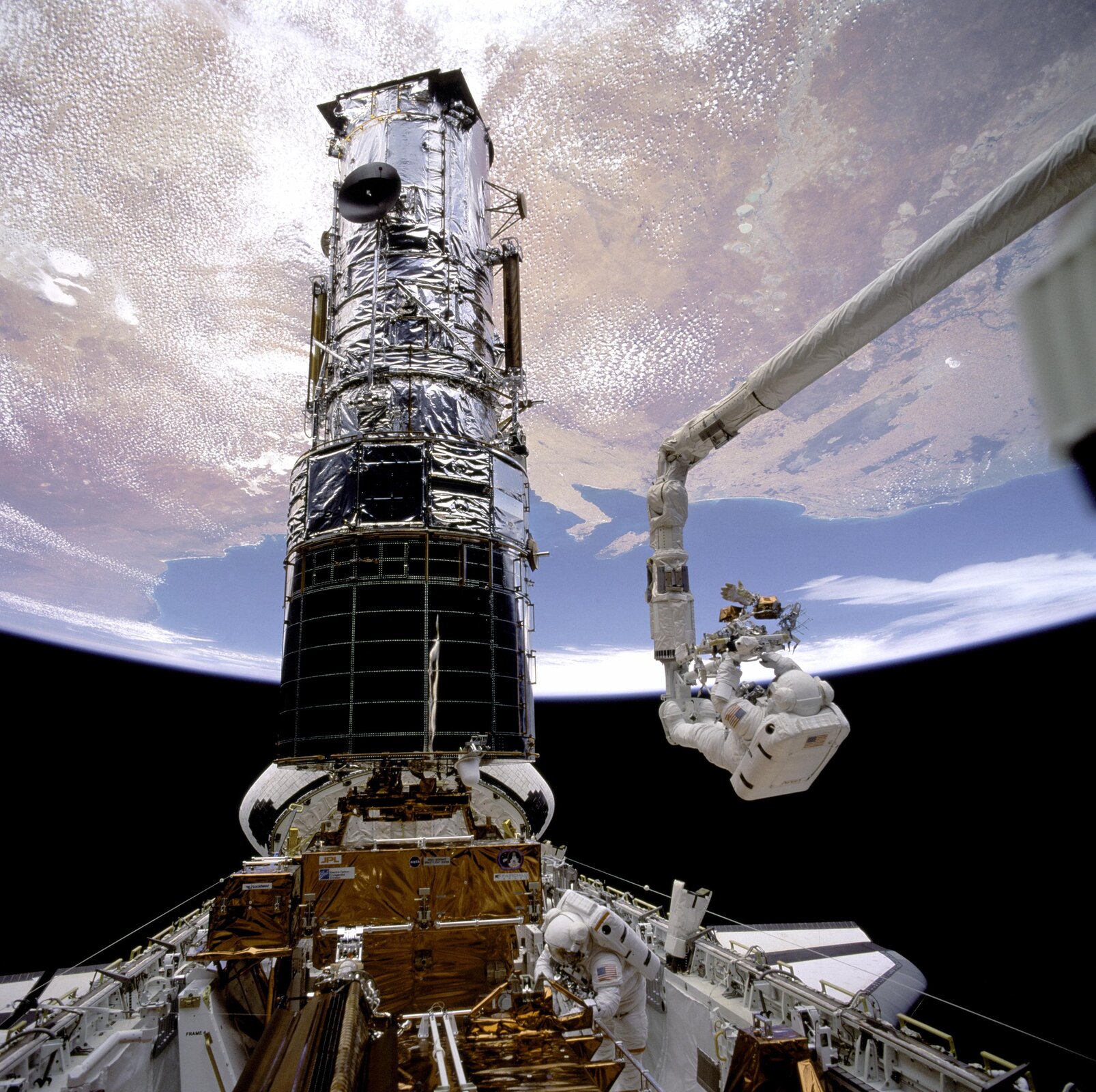 Rys. 1b. Zdjęcie prezentuje teleskop Hubble'a, w ujęciu pionowym. Nad nim unosi się Ziemia. W prezentowanej części teleskopu znajduje się cały jego układ optyczny (zwierciadła) służący do obrazowania danych zarejestrowanych przez urządzenie. Dolna podstawa cylindrycznego kształtu przytwierdzona jest do płaskiej stalowej konstrukcji. Po lewej stronie obok cylindrycznej części teleskopu widoczne jest białe, stalowe ramię wyciągnięte w kierunku teleskopu. Na końcu ramienia zawieszony jest astronauta ubrany w biały kombinezon kosmiczny. Astronauta dokonuje modernizacji teleskopu.