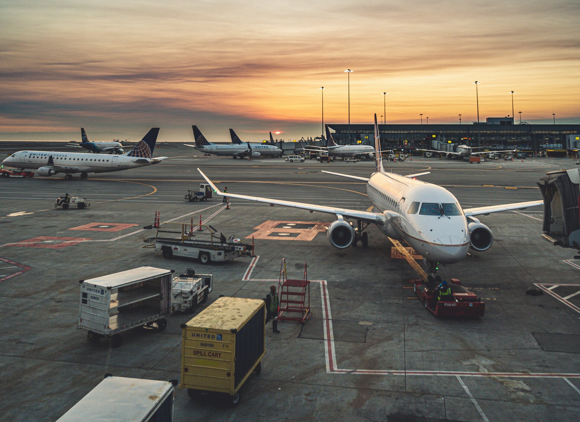 Zdjęcie przedstawia płytę lotniska na której znajdują się samoloty. Widoczne są też wózki i pojazdy do transportu bagaży.