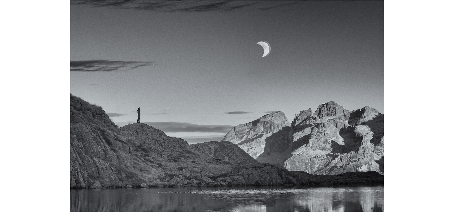 Zdjęcie przedstawia skaliste góry nad zbiornikiem wodnym na tle nieba. Na jednym wzgórzu widnieje postać człowieka stojącego nocą na skale w górach, spoglądającego na Księżyc.