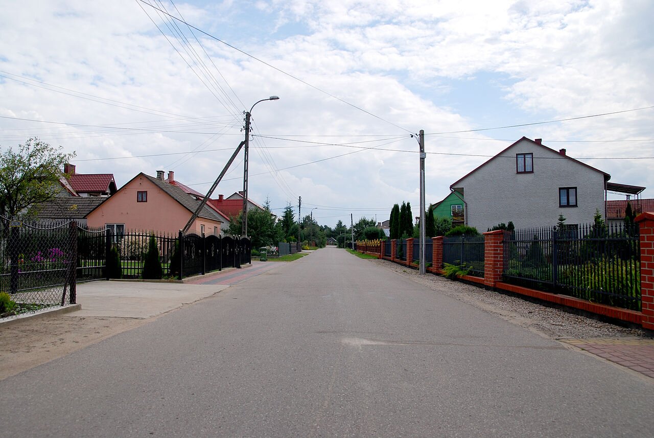 Ilustracja przedstawia wieś w województwie podlaskim na planie tzw. ulicówki. Domy znajdują się po dwóch stronach drogi biegnącej środkiem.