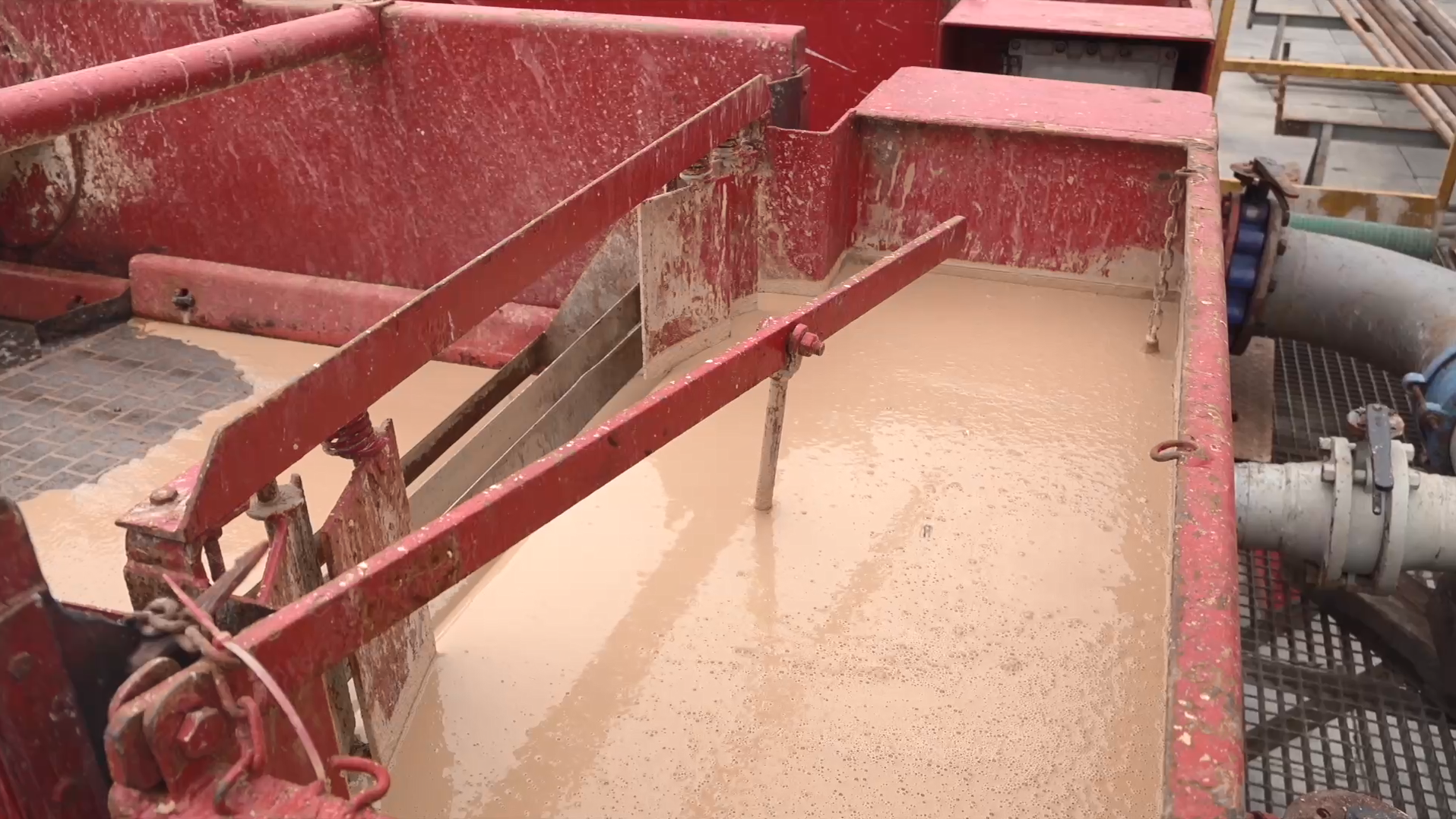 Na zdjęciu widoczny jest zbiornik z  płuczką wiertniczą. Zbiornik jest w kształcie czerwonego prostopadłościanu. W środku zbiornika znajduje się beżowa, gęsta ciecz. Od boku zbiornika z prawej strony odchodzą dwie szare rury. 