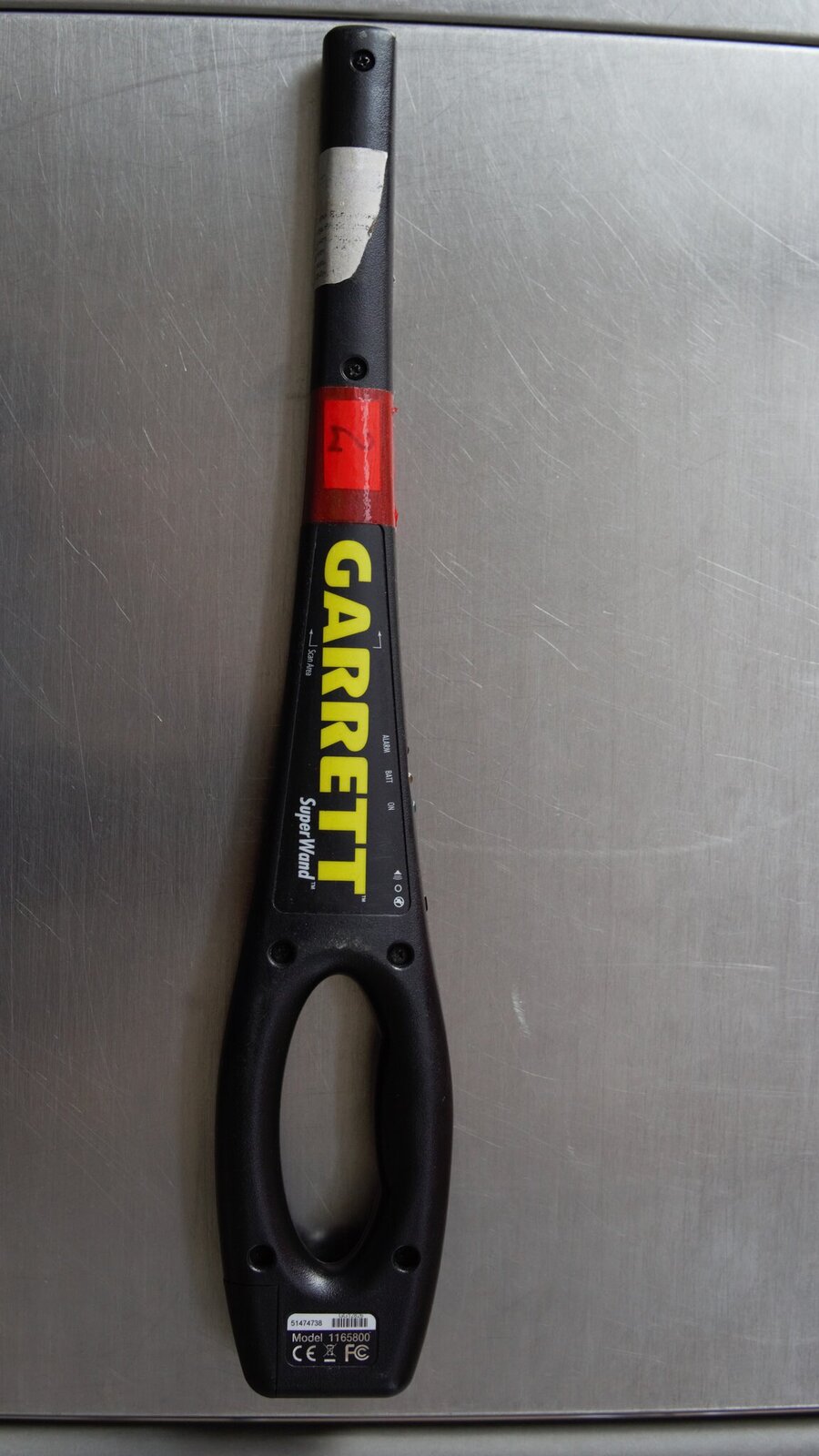 Zdjęcie przedstawia Ha Ha eM De. W nawiase Hend held metal detektor. czyli ręczny wykrywacz metalu do kontroli osób, jako dodatkowe środki kontroli, nie używane samodzielnie. Urządzenie jest czarne. Są na nim napisy. Garet. Super Łond. Ma z tyłu rączkę, a następnie zwęża się w cienki walec. W części walcowatej jest detektor.