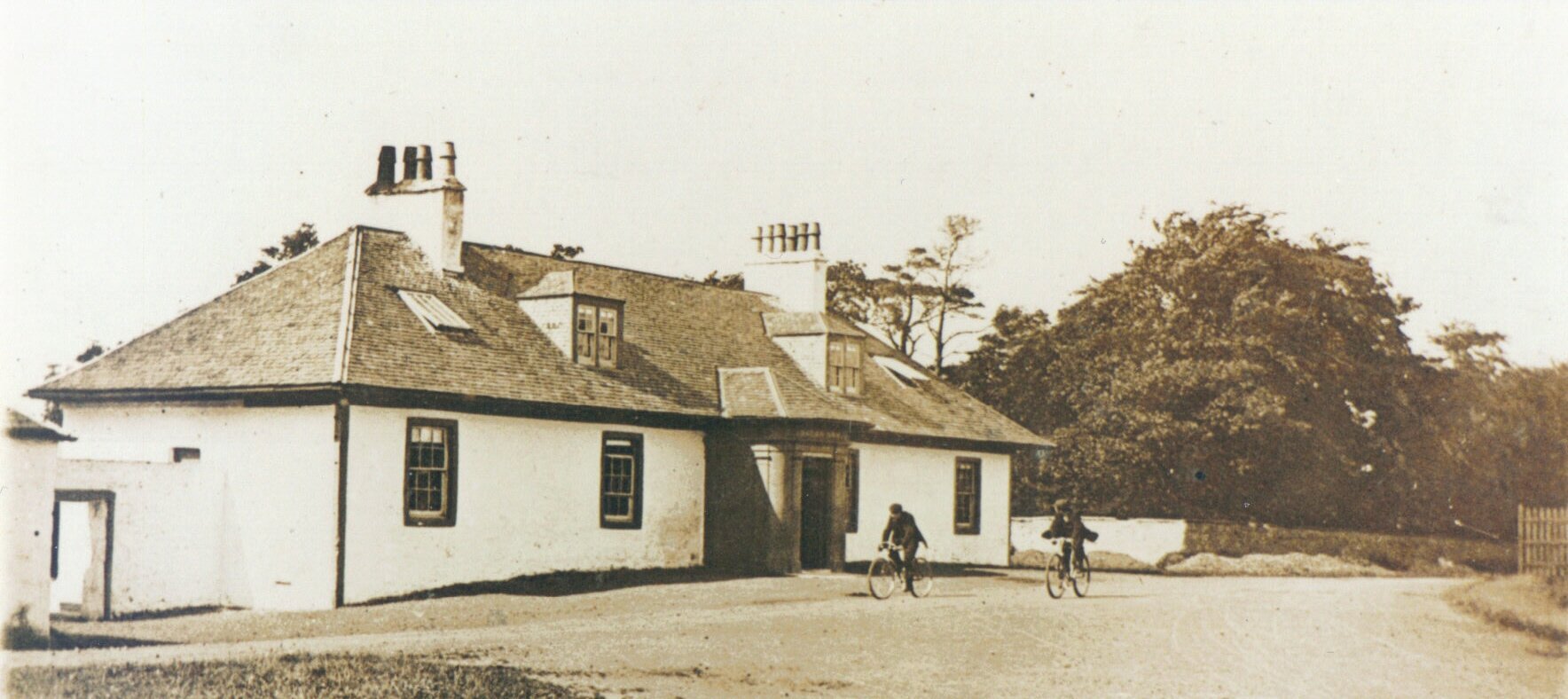 Fotografia czarno-biała przedstawiająca budynek Lugton Inn at Lugton, East Ayrshire,  w Szkocji. Przed domem dwóch rowerzystów. 