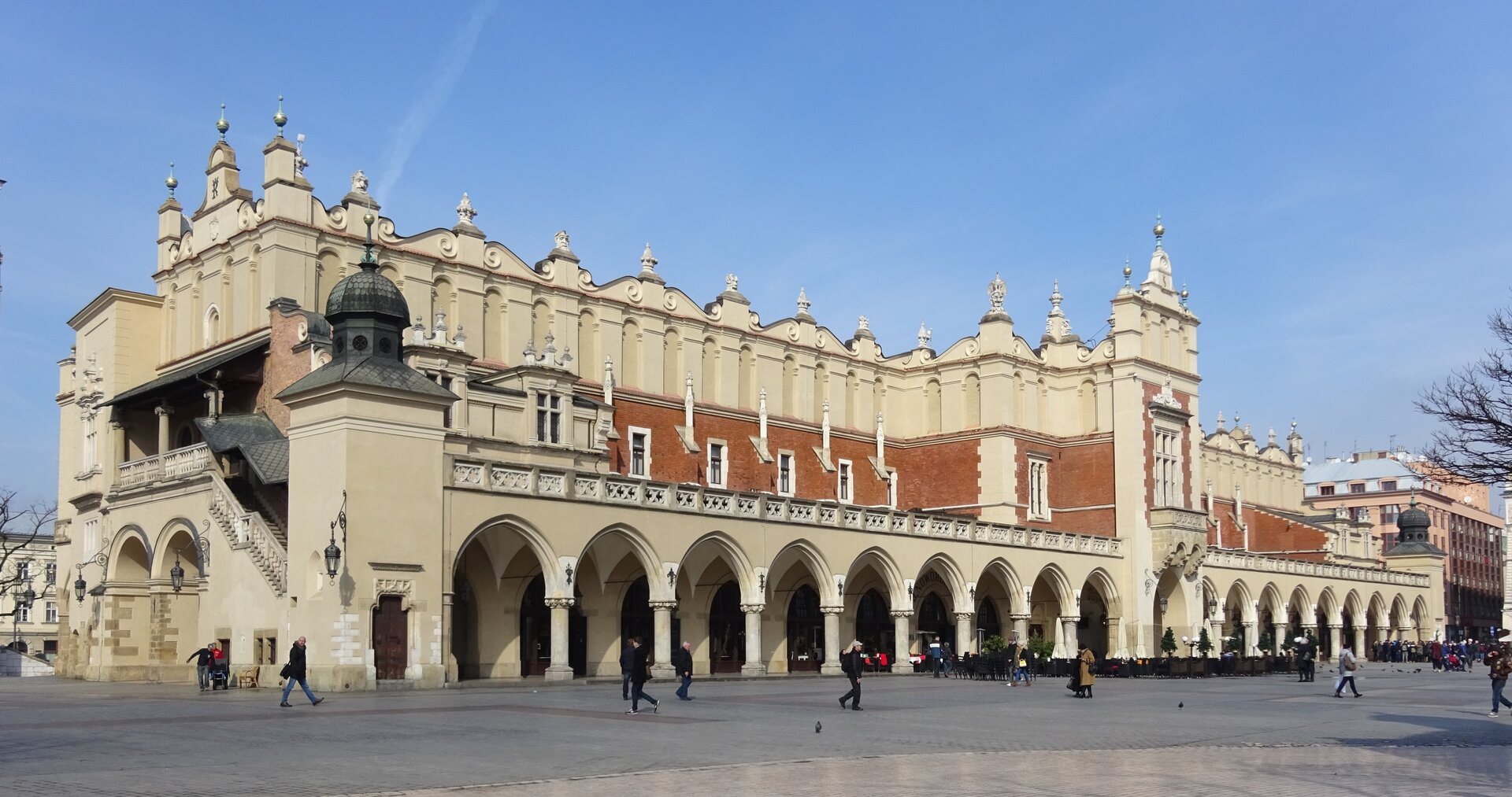 Ilustracja przedstawia Sukiennice w Krakowie, widok z południowego wschodu. Zabytkowy budynek sukiennic znajdujący się w centralnej części Rynku Głównego w Krakowie. Po obu stronach bryły dobudowano arkadowe loggie. Attyka zgodna jest z podziałem arkad. Wieńczą ją maszkarony autorstwa Santi Gucciego.