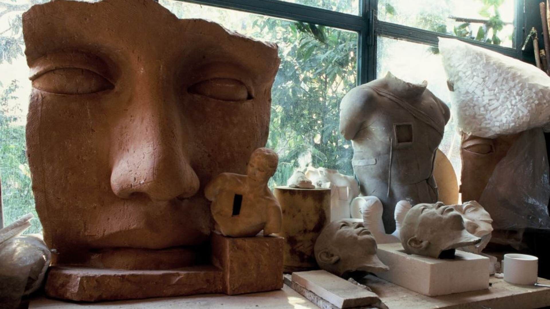 Fotografia przedstawia fragment wnętrza pracowni Igora Mitoraja. Na parapecie, przy oknie stoją różnego rodzaju rzeźby wykonane z różnych materiałów. Pierwsza i największa z rzeźb to Maska - ludzka twarz wykonana z brązowej gliny. Twarz posiada oczy, nos i usta, nie przedstawia żadnych emocji. Pod prawym okiem rozmieszczone są białe punkciki. Następnie, z prawej strony leży popiersie mężczyzny, który opiera się na prawym amputowanym ramieniu. Twarz mężczyzny nie wzbudza żadnych emocji. Popiersie leży na brązowym podeście. Obok leżą trzy wyrzeźbione głowy starszych, łysych mężczyzn. W tle znajduje się wyrzeźbiony tułów przepasany materiałem. Za oknem obecne są drzewa oraz krzewy.
