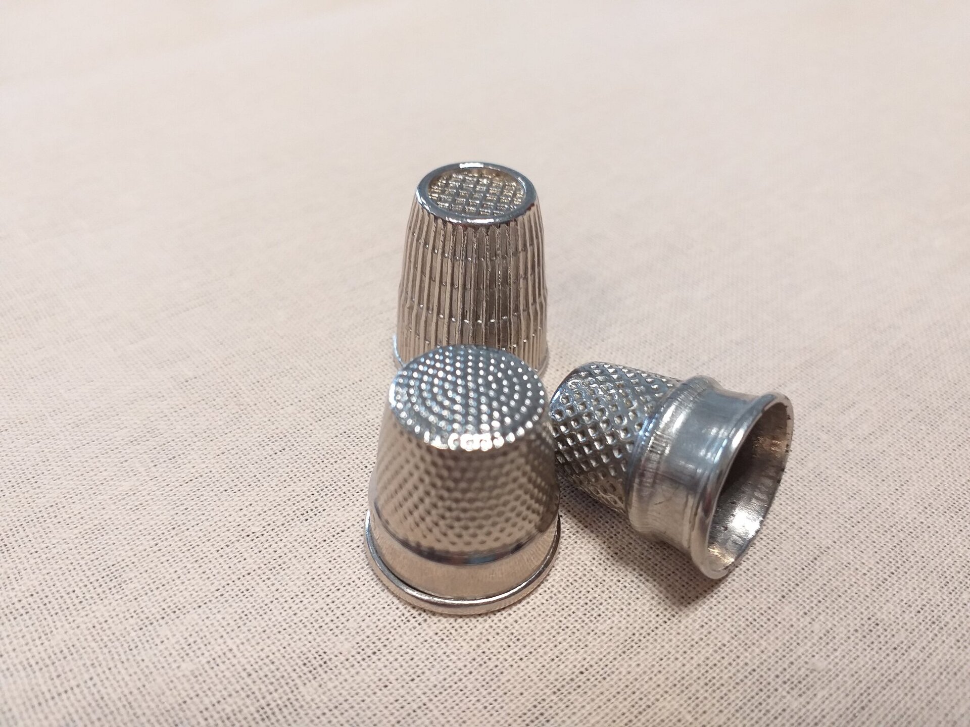 Zdjęcie przedstawia trzy metalowe naparstki leżące na płótnie. Naparstki są kształtem dostosowane do kształtu palca wskazującego ręki.