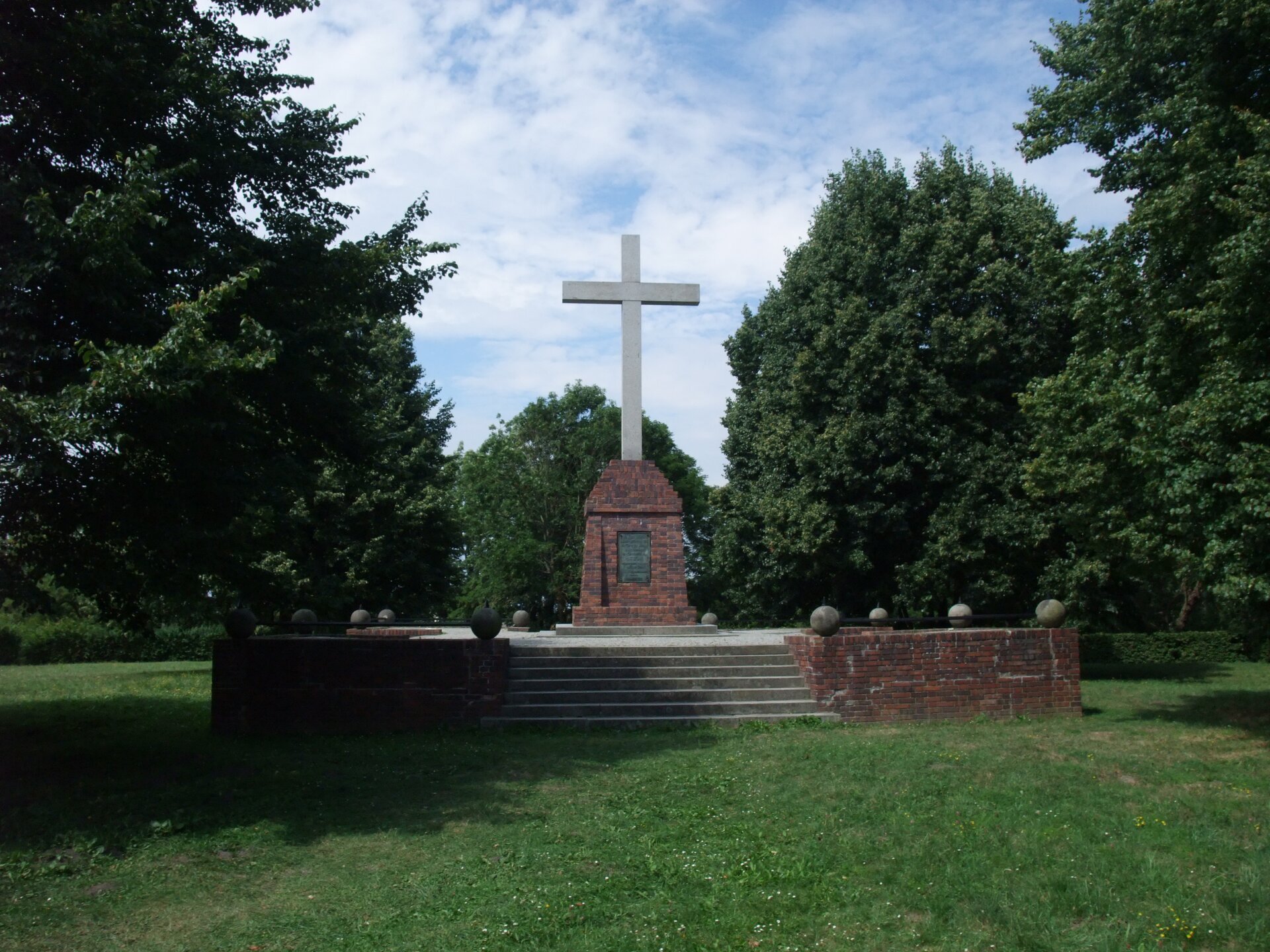 Widok na cokół ceglany z krzyżem kamiennym pośrodku. Na pomnik prowadzi postument ze schodami, z zewnątrz wyłożony cegłą, widoczne są także kamienne kule, rozłożone po obwodzie zewnętrznego okręgu. Widoczna tablica pod kamiennym krzyżem. W tle polana i zagajnik. Zdjęcie współczesne.