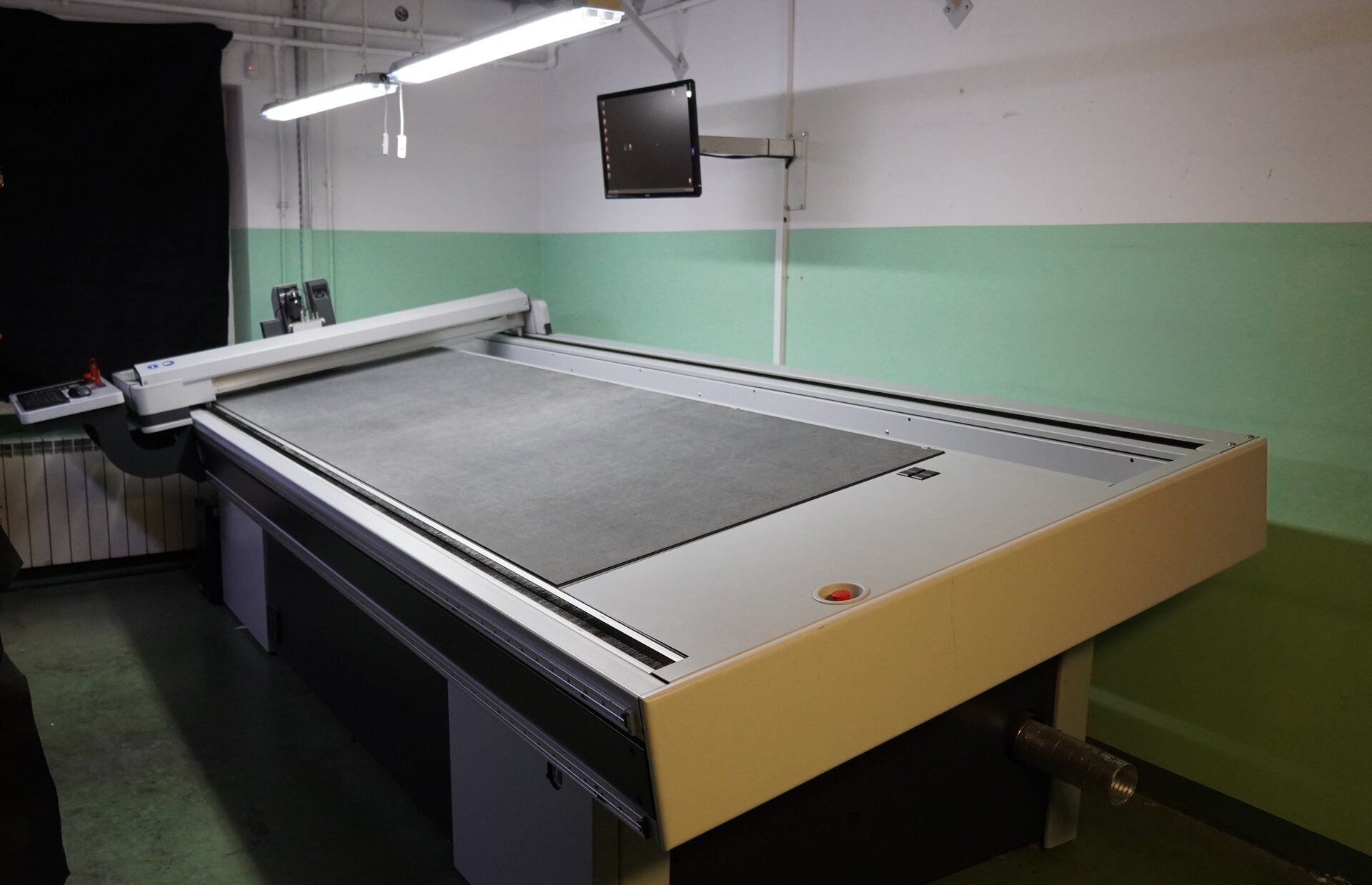 Zdjęcie przedstawia duży pulpit, na którym znajduje się kawałek materiału. Po lewej stronie znajduje się narzędzie, które umożliwia rysowanie na materiale za pomocą komputera. Monitor jest zawieszony nad pulpitem. Znajduje się tam także podłużna lampa, która oświetla warsztat pracy. Nagranie tożsame z treścią.