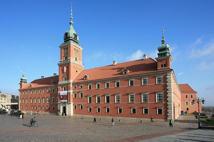 Fotografia barwna przedstawia zamek królewski w Warszawie od strony Placu Zamkowego. Ma kształt zamkniętego pięcioboku. Na fotografii widać tylko jedną, dwupiętrową ścianę budynku. Po jego bokach znajdują się dwie wieże, zwieńczone ostrymi szpikulcami. Trzecia z zegarem, największa znajduje się na środku budynku, pod wejściem na dziedziniec zamkowy. Okna są prostokątne, mniejsze na parterze, największe na pierwszym piętrze. Przed budynkiem znajduje się plac wyłożony kamiennymi płytami, po którym spaceruje kilkoro ludzi.