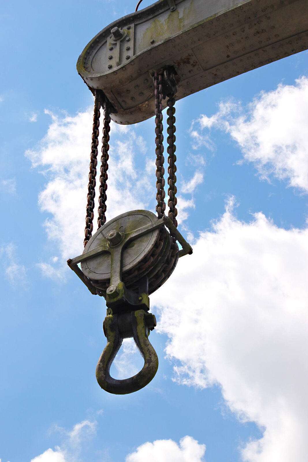 Zdjęcie przedstawia elektryczną dźwignię. Dźwignia ma postać prostopadłościennego urządzenia z zawieszonym na stalowej lince hakiem. Linka nawinięta jest na okrągły bloczek, widoczny w dolnej części urządzenia i zawieszony na dwóch stalowych linach. Wewnątrz prostopadłościennej części urządzenia zamontowany jest silnik elektryczny, który wykonując pracę nawija lub rozwija stalową linkę, na której zawieszony jest hak. 