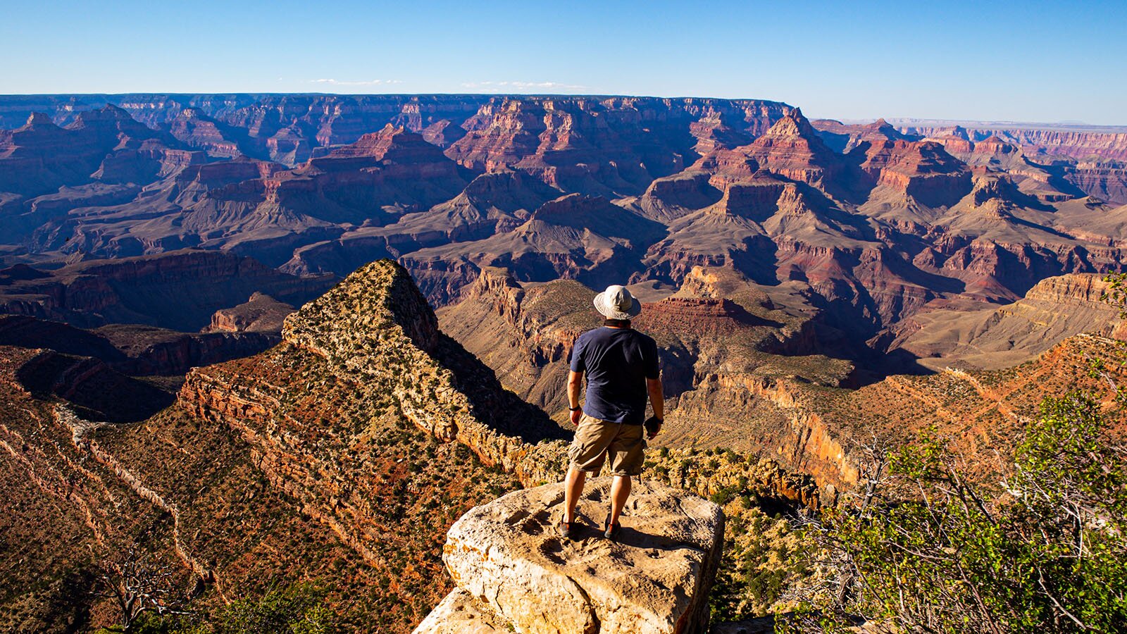 Zdjęcie przedstawia mężczyznę, w ujęciu od tyłu, stojącego na płaskiej skale i patrzącego na skaliste łańcuchy kanionu. Mężczyzna jest ubrany w granatową koszulkę z krótkim rękawem, jasne szorty, a na głowie ma jasny kapelusz. Przed nim rozciągają się pasma skalistych formacji w kolorze brązowo‑czerwonym. Na horyzoncie widoczny jest skalisty płaskowyż.