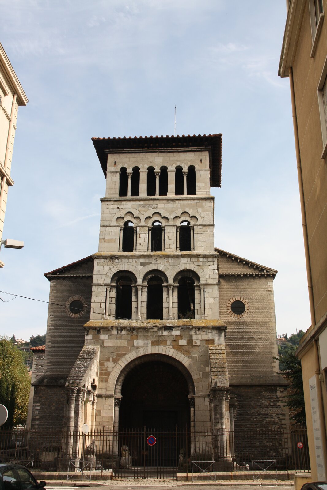 Zdjęcie przedstawia widok współczesny na fasadę kościoła pw. św. Marcina w Vienne we Francji. Fasada jest jednocześnie wysoką wieżą. Na parterze znajduje się duże wejście, wokół którego znajduje się romański portal. Powyżej dwa rzędy portali: pierwszy o łukach pełnych, drugi trójlistny. Otwory okienne przedzielone są kolumnami. Na szczycie wieży widoczne trzy otwory o charakterze portalu rozdzielone kolumną. Na szczycie wieży płaski dach. Po obu bokach wieży widoczne ściany zewnętrzne z okrągłymi, małymi oknami oraz skośnym dachem. Budynek oddzielony od ulicy metalowym płotem, widoczny na bramie znak zakazu zatrzymania. Przy obu krawędziach zdjęcia fragmenty sąsiadujących z kościołem budynków. Niebo pogodne. W tle zieleń - korony drzew.