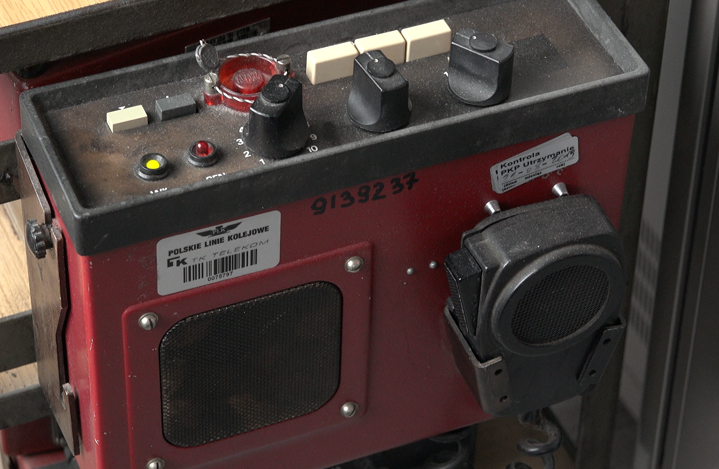 Zdjęcie pokazuje radiostację łączności bezprzewodowej. Jest to czerwona metalowa skrzynka. Na jej największym boku widoczny jest głośnik oraz zawieszony mikrofon ręczny z przyciskiem do nadawania, połączony ze skrzynką kablem. Na górnej podstawie skrzynki znajduje się kilka białych przycisków i jeden szary, dwie lampki, zielona i czerwona, oraz trzy pokrętła. Jest tam również jeden większy okrągły czerwony przycisk z napisem Alarm.
