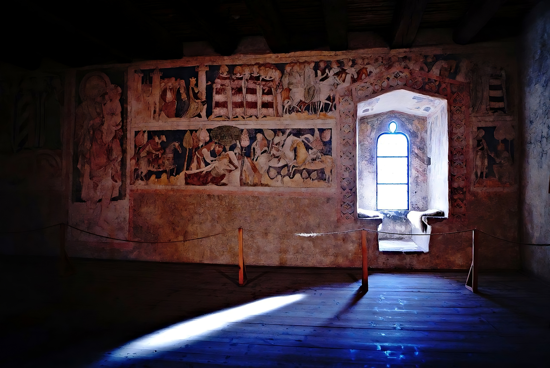 Fotografia przedstawia wnętrze ciemnego pomieszczenia. Z prawej strony widoczna jest głęboka wnęka z wysokim, wąskim oknem. Z lewej strony, obok okna, na całej ścianie, malowidła ułożone w dwóch, poziomych rzędach. Na malowidłach liczne sylwetki mężczyzn, kobiet oraz zwierząt.