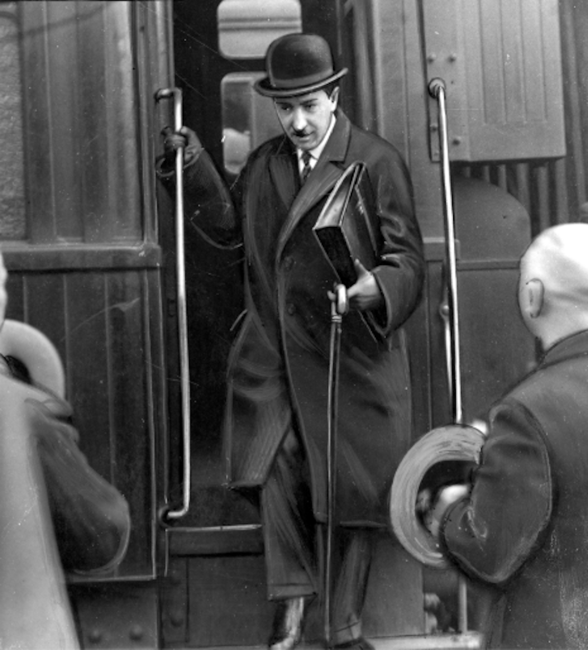 Ilustracja przedstawia mężczyznę  wysiadającego z pociągu. Mężczyzn ubrany jest w cylinder, płaszcz oraz garnitur wystający spod płaszcza. Na dłoniach ma skórzane rękawiczki. W prawej ręce trzyma aktówkę oraz laskę.  Przed nim widać kontury dwóch innych osób zwróconych do niego twarzą.