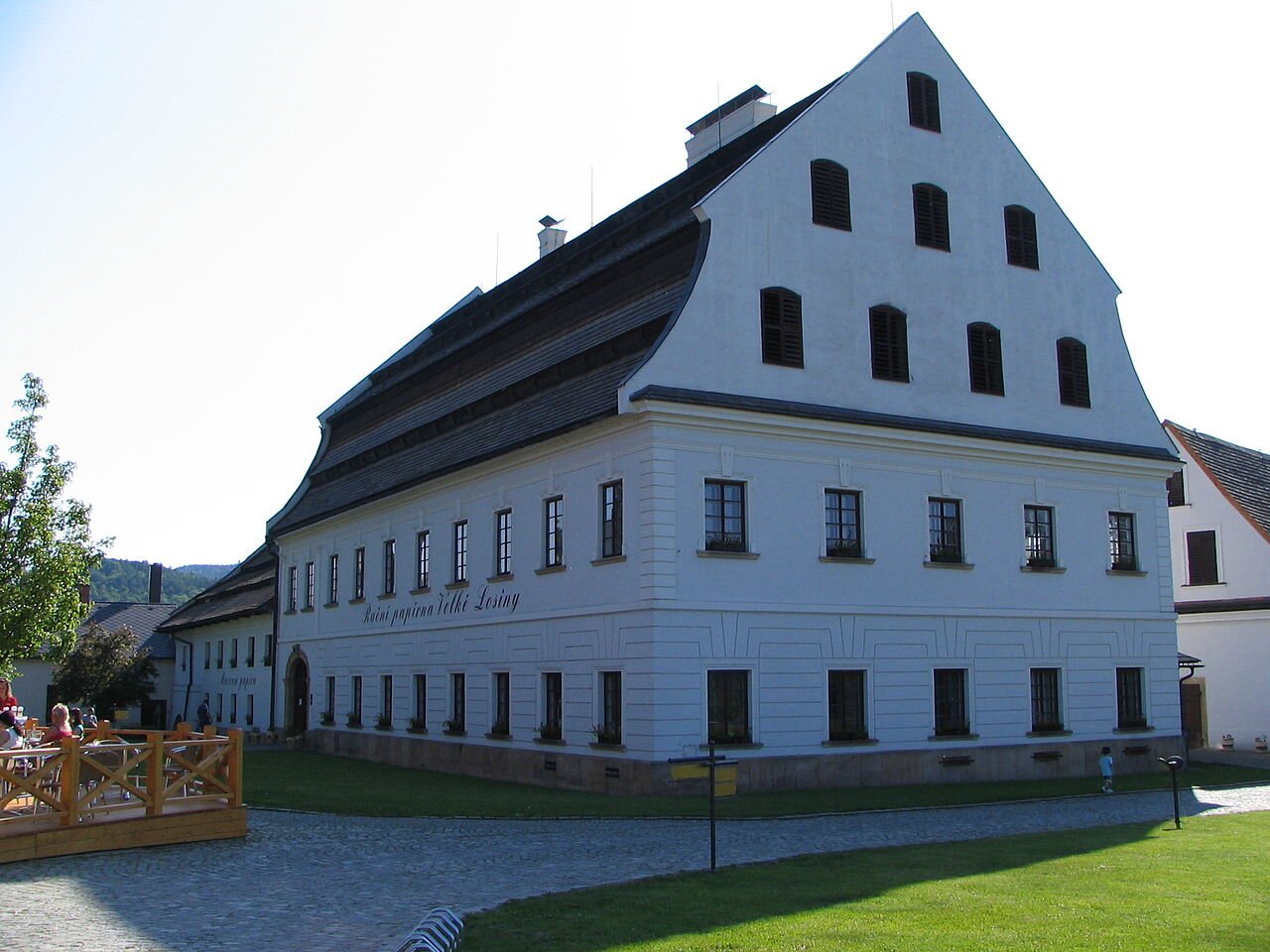 Zdjęcie starego budynku o białej, surowej fasadzie, na której znajdują się rzędy okien. Duże poddasze. Dookoła chodnik, trawa. W tle mniejsze budynki.