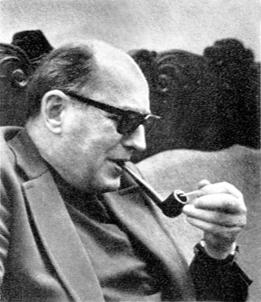 Zdjęcie przedstawia dojrzałego, tęgiego mężczyznę ukazanego prawym profilem. Mężczyzna jest łysiejący, nosi okulary. Trzyma w ustach fajkę, którą zapala lewą ręką. Mężczyzna ubrany jest w garnitur.