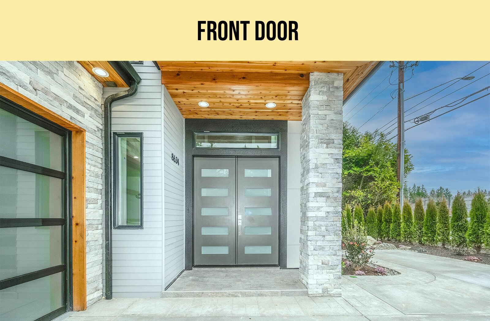 Zdjęcie przedstawia dwuskrzydłowe, masywne drzwi wejściowe do domu. Budynek zbudowany jest z szarego kamienia i drewna. Nad wejściem palą się dwie lampki sufitowe. Po lewej stronie widoczne jest duże okno. Na prawo od budynku widoczny jest fragment chodnika, rząd tui oraz linia energetyczna. U góry widoczny jest napis w języku angielskim: FRONT DOOR.