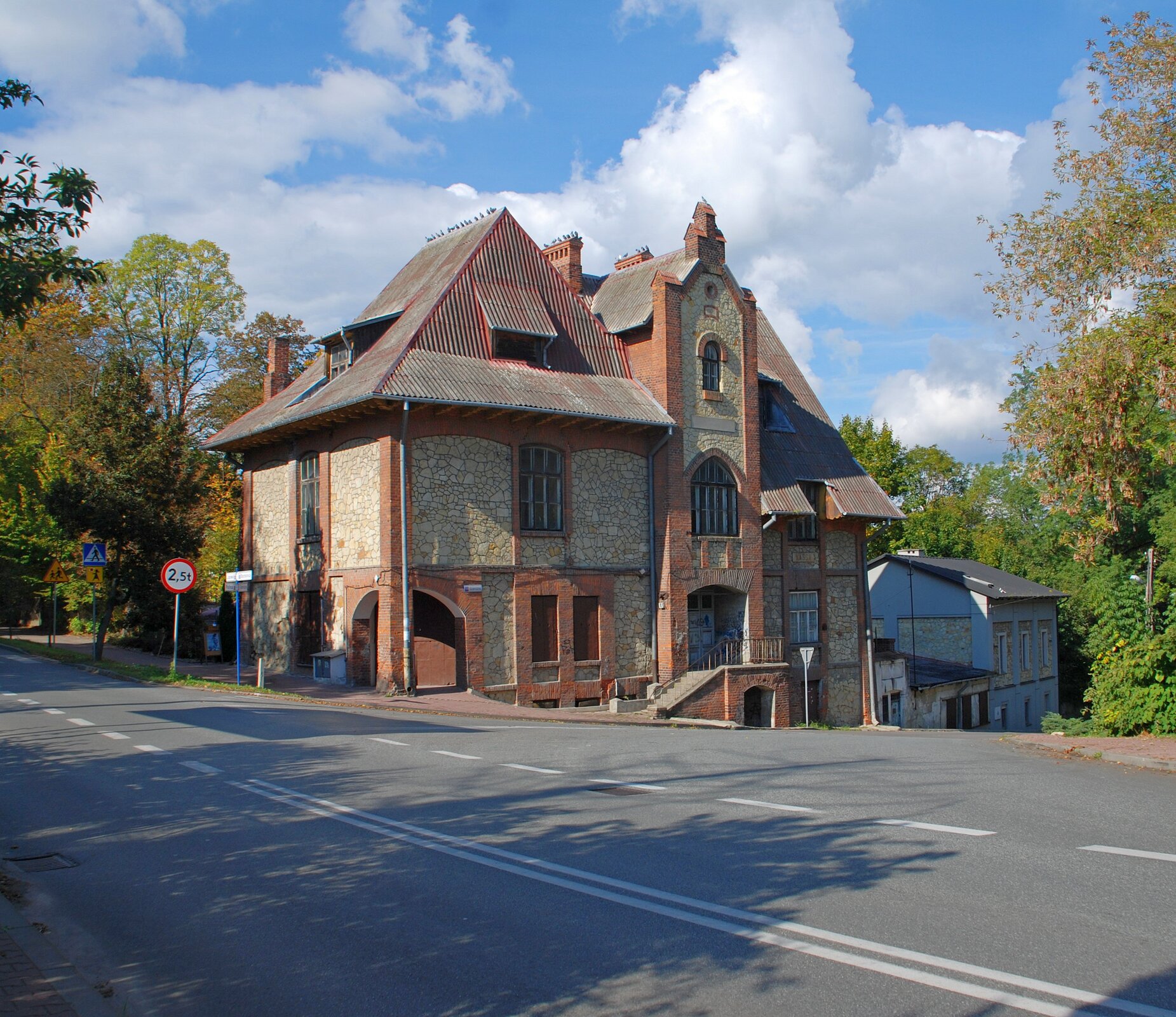 Zdjęcie przedstawia dom stojący przy ulicy, którego ściany zewnętrzne wykonane są z równo ułożonego kamienia. Dom ma spadziste dachy. Wybudowany jest na pochyłej działce.