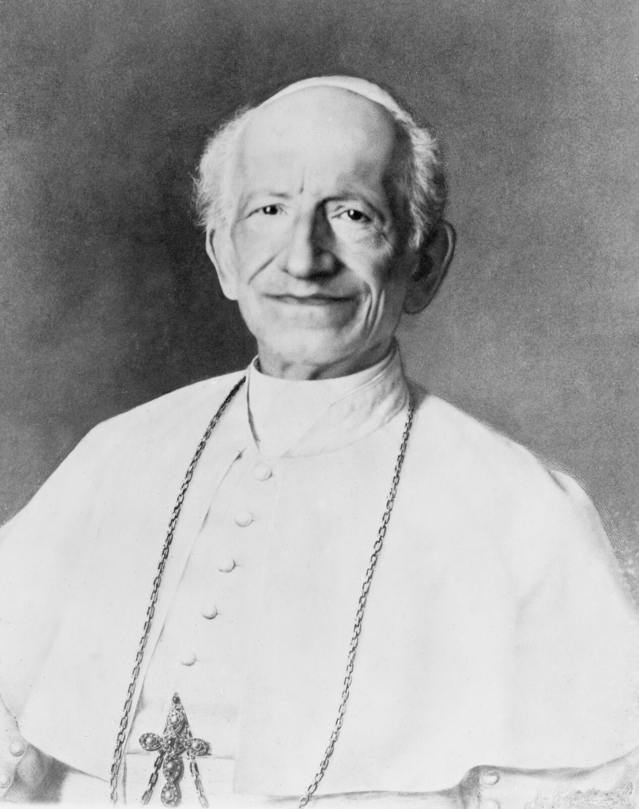 Zdjęcie przedstawia starszego mężczyznę w białym, papieskim stroju i piusce na głowie. Na jego szyi wisi krzyż na długim łańcuchu.