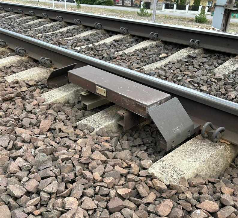 Zdjęcie przedstawia fragment torowiska. Po zewnętrznej stronie prawej szyny widoczne jest prostopadłościenne urządzenie przymocowane do szyny, po jego bokach znajdują się dwie wygięte blachy. Urządzenie to nazywa się rezonator torowy. Jest to element wchodzący w skład samoczynnego hamowania pociągu. 