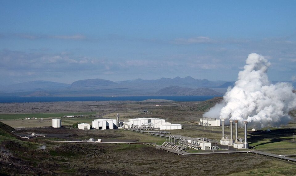 Ilustracja przedstawia elektrownię geotermiczną w Islandii. Z czterech kominów wydobywa się biały dym. Obok znajdują się budynki przemysłowe, od których odchodzą wielkie rury. W oddali znajdują się góry.