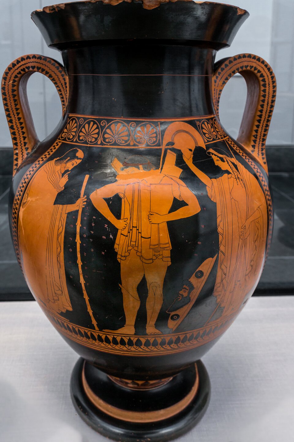 Ilustracja przedstawia grecką wazę czerwonofigurową. Scena ukazuje Hektora uakzuje księcia trojańskiego Hektora w trakcie przygotowań do walki. W centrum kompozycji stoi nagi młodzieniec (Hektor), który zakłada zbroję. Towarzyszą mu dwie postacie. Po lewej znajduje się Priam, jego ojciec, ukazany jako starzec z siwą brodą i laską, ubrany w himation. Prawą ręką udziela błogosławieństwa. Po prawej jest Hekabe - matka Hektora,  trzymająca hełm nad jego głową. 