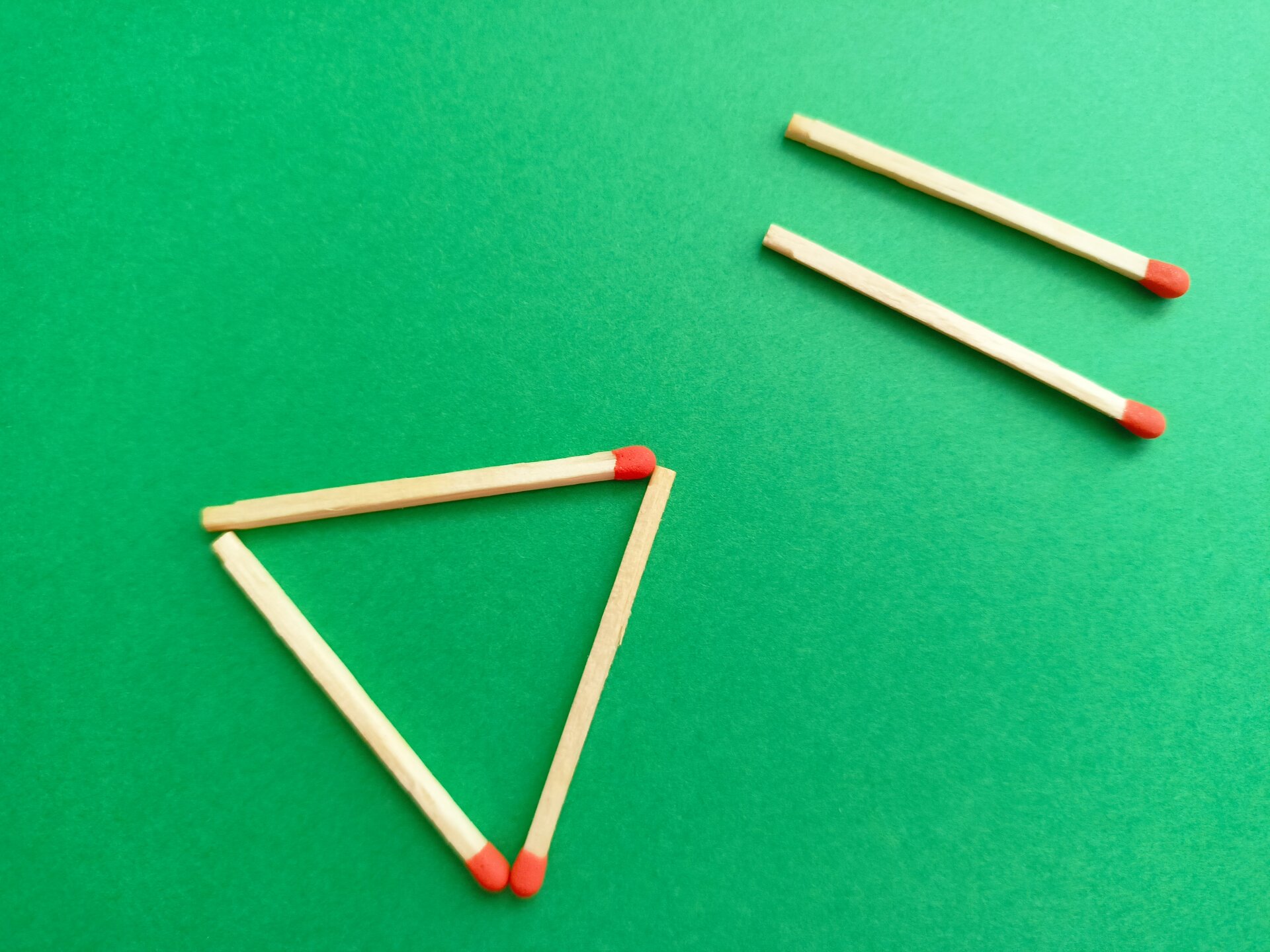 Zdjęcie przedstawia zielony blat, na którym leży pięć zapałek. Dwie są ułożone po prawej stronie, jedna przy drugiej. Trzy są ułożone po lewej stronie, tworzą trójkąt.