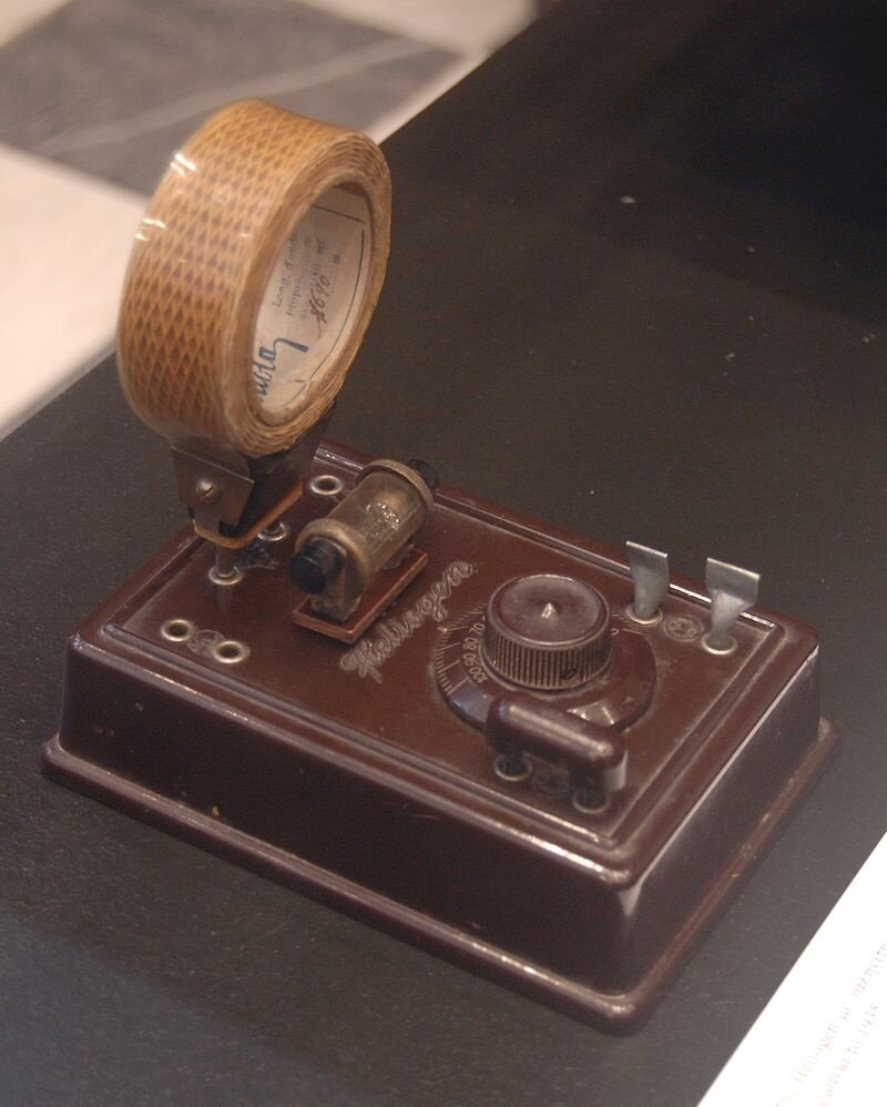 Na zdjęciu znajduje się urządzenie. Jest to płaska skrzyneczka, na której znajduje się jedno duże pokrętło, dwa  małe wystające bolce, jeden poziomy wystający drążek, mała przyczepiona poziomo fiolka oraz tuż przy fiolce wystające ku górze kowadełko, w którym umieszczono rolkę z materiałem o strukturze siatkowanej. Rolka owinięta jest przezroczystą osłonką.