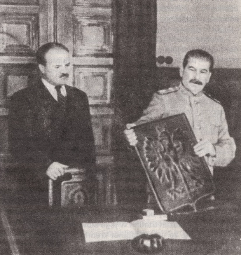 Kliknij, aby powiększyć Na zdjęciu sala, gdzie na tle dużych drzwi stoi za biurkiem dwóch mężczyzn. Mężczyzna po prawej - Józef Stalin - ubrany jest w wojskową marynarkę, prezentuje tablicę z godłem polskim. Na godle jest orzeł bez korony. Mężczyzna po lewej - Wieczysław Mołotow - stoi z dłonią położoną na oparciu fotela.
