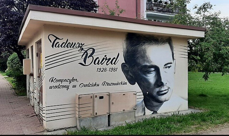 Na ścianie małego budynku znajduje się czarno‑biały mural przedstawiający portret Tadeusz Baird (1928–1981). Obok widnieje napis informujący, że był kompozytorem urodzonym w Grodzisku Mazowieckim; mural znajduje się przy chodniku, w otoczeniu trawy i drzew.