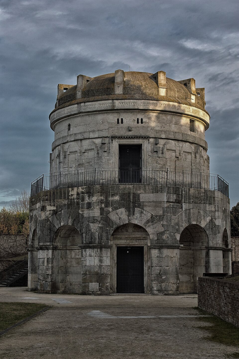 Fotografia przedstawia okrągły budynek z jednym piętrem i kopułą na dachu. Na parterze wokół budynku widać kamienne łuki, w jednym z nich widać drzwi wejściowe do budynku. Całe piętro jest otoczone metalową barierką, po środku są drzwi. Nad budynkiem widać zachmurzone niebo.