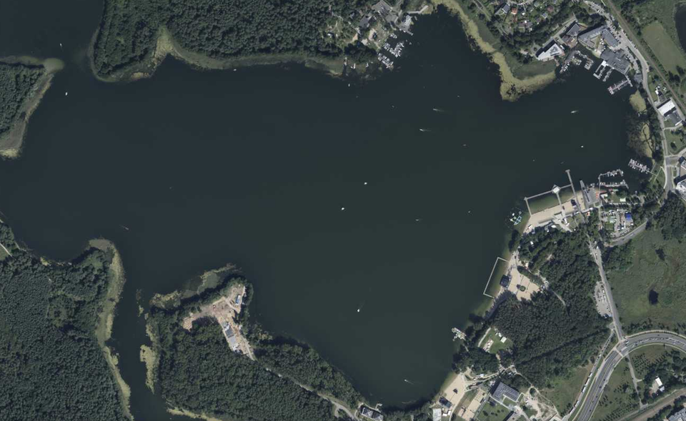 Zdjęcie satelitarne przedstawia fragment jeziora Ukiel w Olsztynie. Jezioro ma zaokrąglone brzegi. Na brzegu znajduje się molo, plaże oraz budynki.