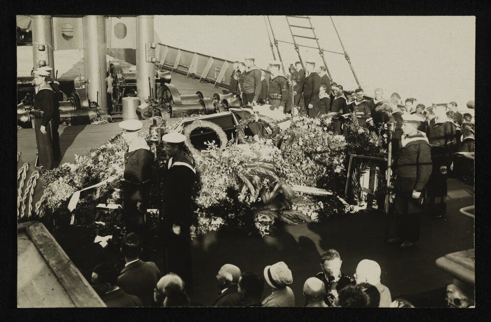 Zdjęcie przedstawia pokład statku. Są na nim zgromadzeni licznie ludzie. Na środku pokładu stoi trumna. Jest obłożona wieloma wieńcami kwiatów. Wokół trumny pełnią wartę marynarze. Wokół nich widać także kobiety i mężczyzn.