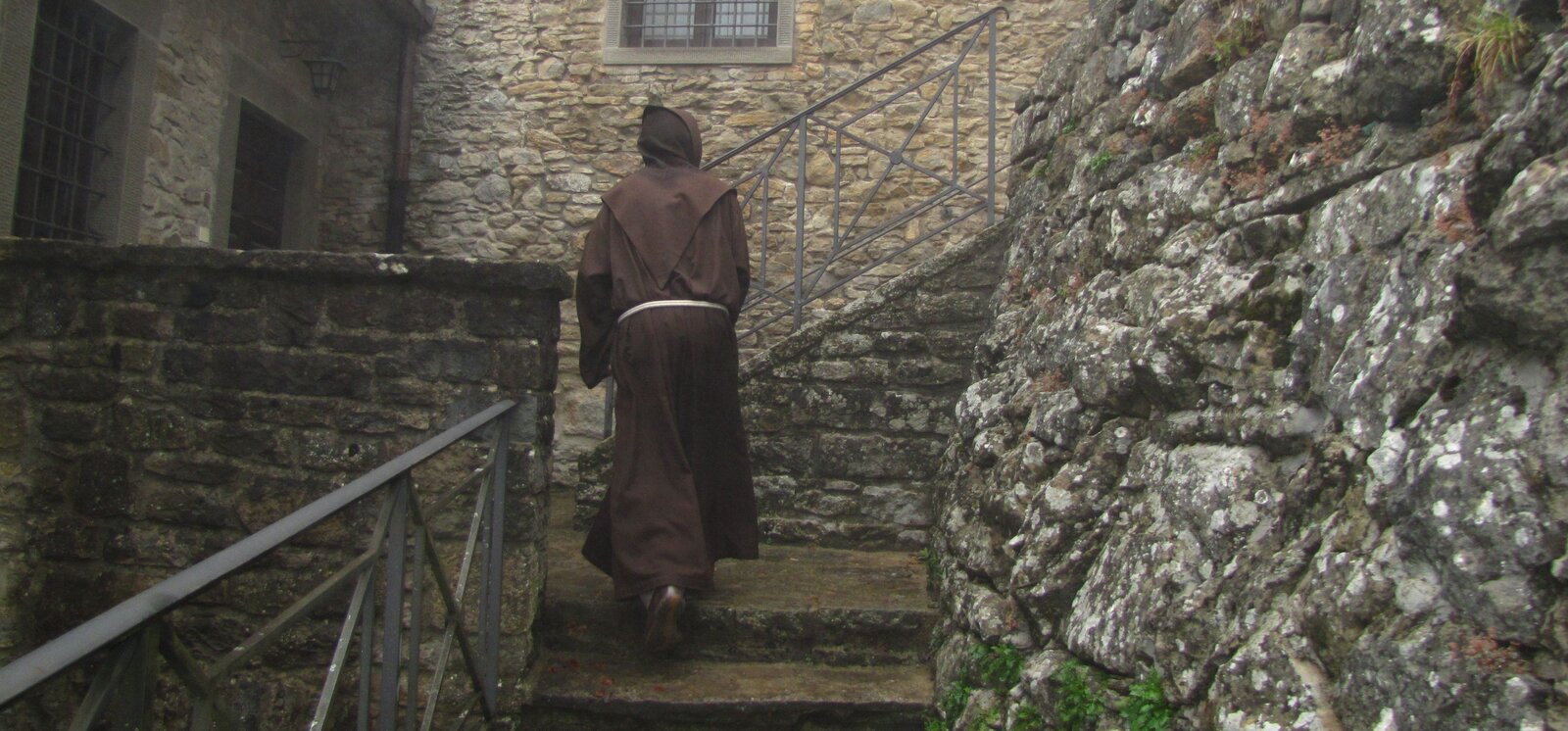 Mnich wchodzi po schodach do klasztoru z kamienia. Jest w brązowym habicie. Na głowie ma kaptur i biały sznur w pasie. Mnich jest odwrócony tyłem. 