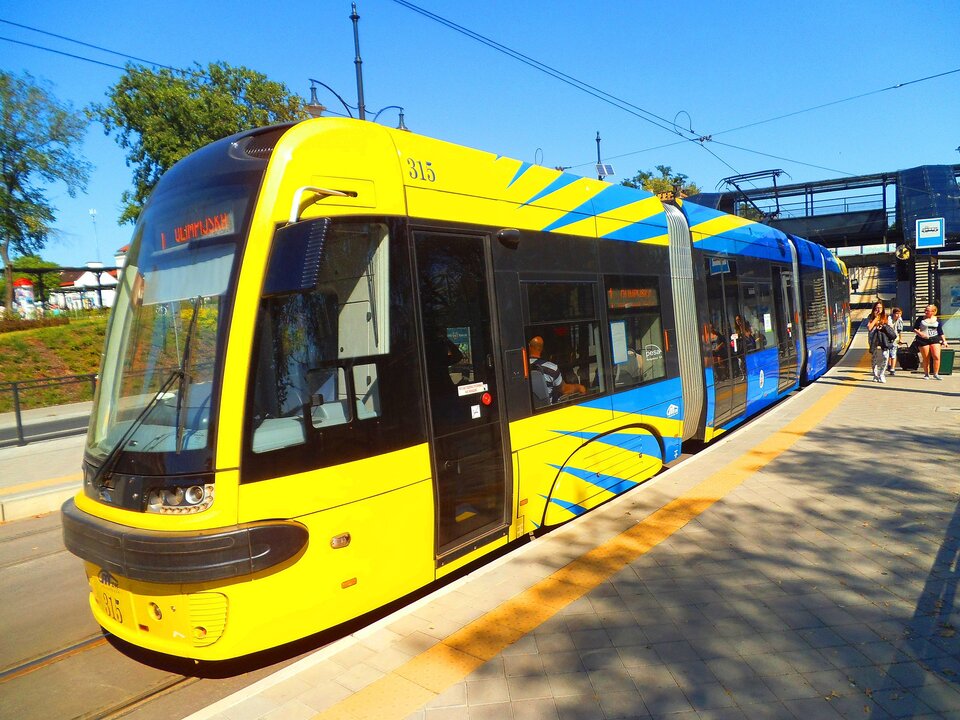 Zdjęcie przedstawia nowoczesny tramwaj, niskopodłogowy, stojący na przystanku tramwajowym. Na stacji jest kilka osób. 