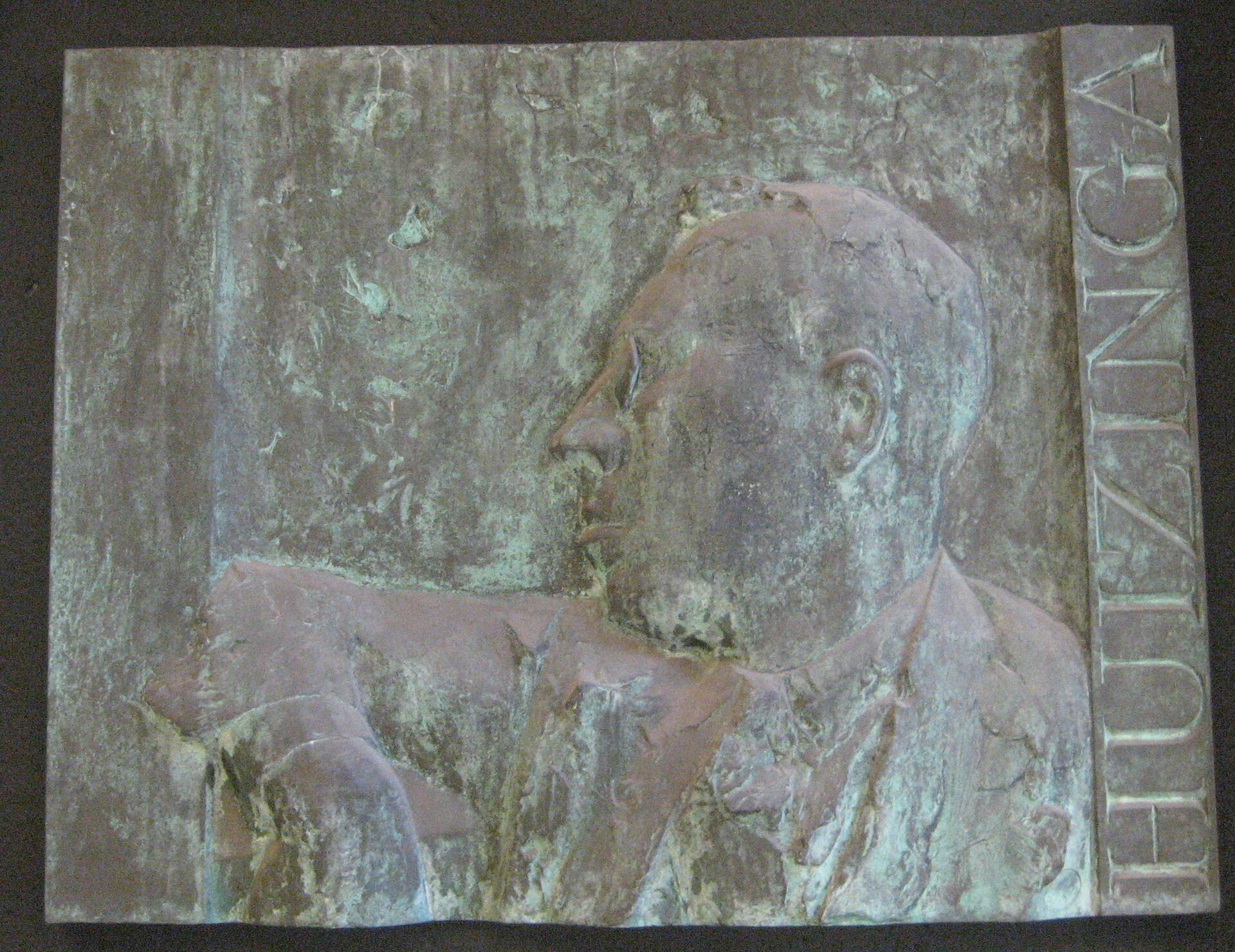 Zdjęcie przedstawia tablicę z płaskorzeźbą głowy mężczyzny. Ukazany jest jego lewy profil i napis po prawej stronie: Huizinga. Płaskorzeźba jest niewyraźna.