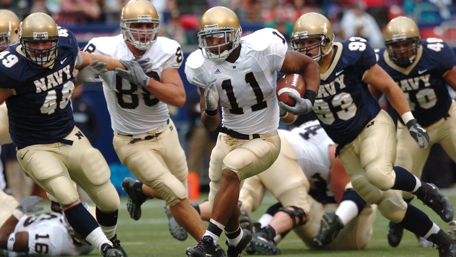 Zdjęcie przedstawia zawodników dwóch drużyn futbolu amerykańskiego. Mężczyźni są w ruchu, biegnąc odpychają przeciwników. Mają na sobie kaski z ochraniaczami na twarz, koszulki z numerami, pod którymi odciskają się ochraniacze na barki oraz klatkę piersiową. Ich spodenki sięgające kolan, wyposażone są również w ochraniacze.