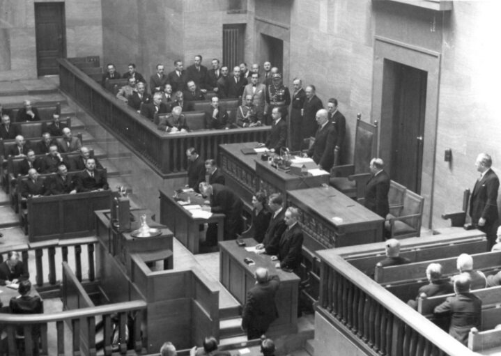 Zdjęcie przedstawia salę sejmową. Marszałek sejmu stoi za stołem i przemawia do zebranych posłów. 