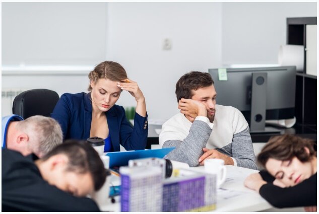 Ilustracja przedstawia zdjęcie znużonych osób podczas firmowego zebrania. Część osób śpi trzymając głowy oparte na blacie biurka. Jeden z mężczyzn podpiera głowę prawą ręką i patrzy gdzieś przed siebie. Obok niego siedzi skupiona kobieta. Na stole stoją kubki, leżą papiery i inne dokumenty firmowe. 