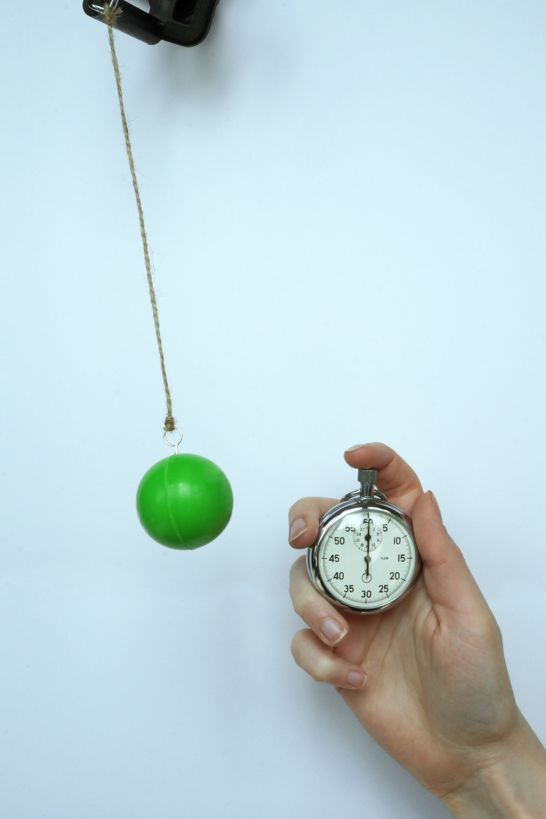 Zdjęcia wahadła matematycznego i stopera trzymanego przez dłoń. Zielona kula wahadła zawieszona jest na sznurku i znajduje się w wychylonej pozycji. Stoper wskazuje 0 s. Tło jasnoniebieskie.