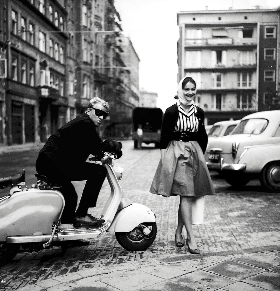 Zdjęcie przedstawia parę młodych ludzi z lat 60-tych. Mężczyzna siedzi na skuterze w przeciwsłonecznych okularach i rękawicach, odwraca głowę w kierunku fotografującego. Obok stoi dziewczyna w rozłożystej spódnicy do kolan i chustce na głowie, uśmiecha się.