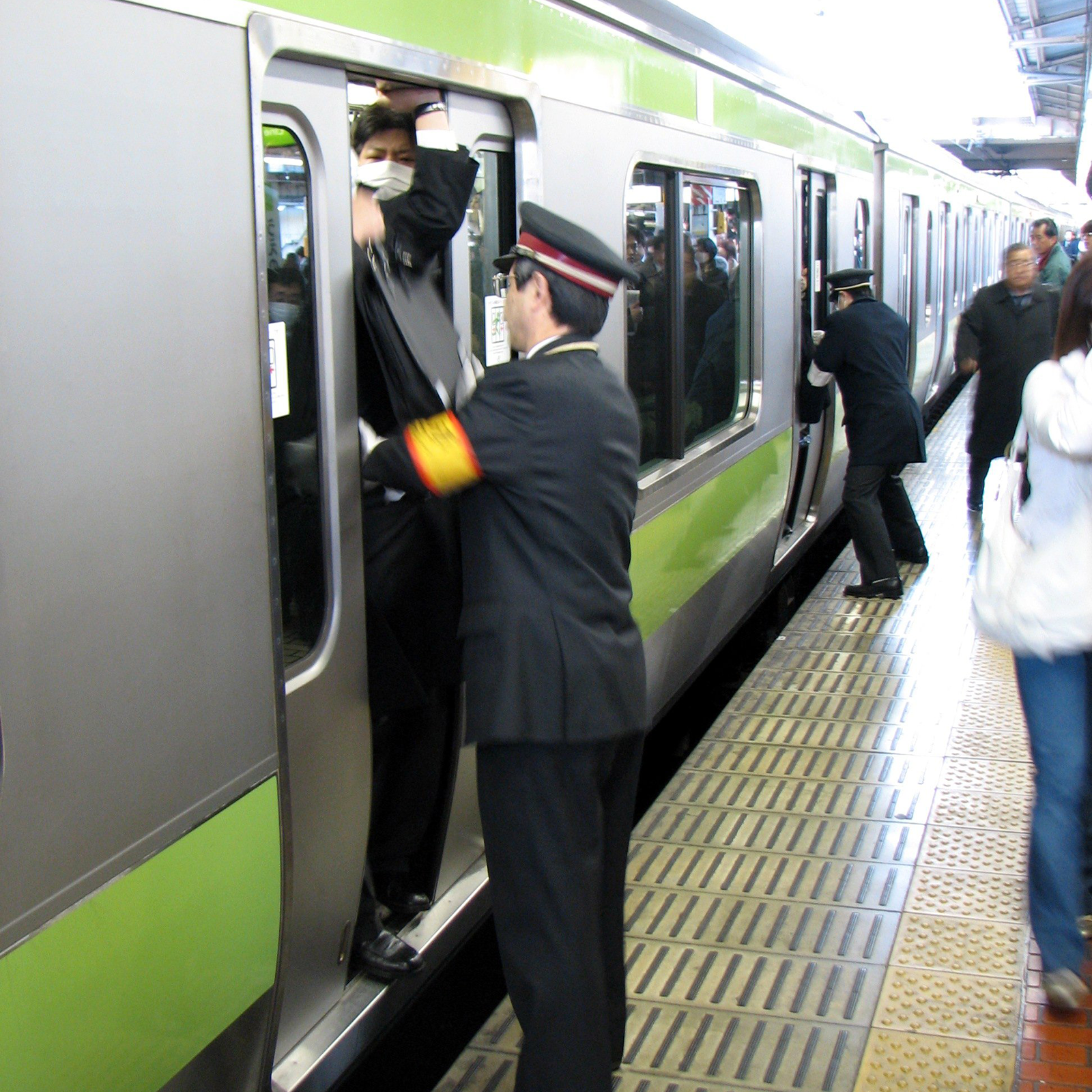 Zdjęcie przedstawia pociąg stojący przy peronie. Na peronie przy drzwiach pociągu stoi dwóch mężczyzn. Jeden z nich trzyma ręce na osobie, która częściowo wystaje poza drzwi.
